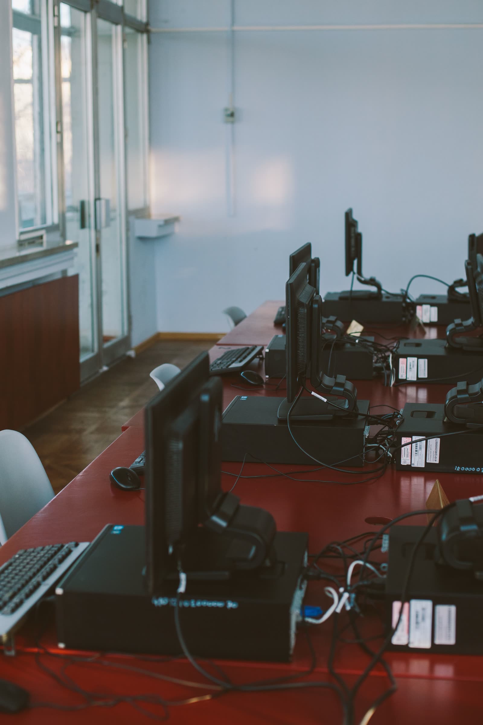 A daylit computer room with monitors and towers lined along a bright window wall, no coloured fixtures